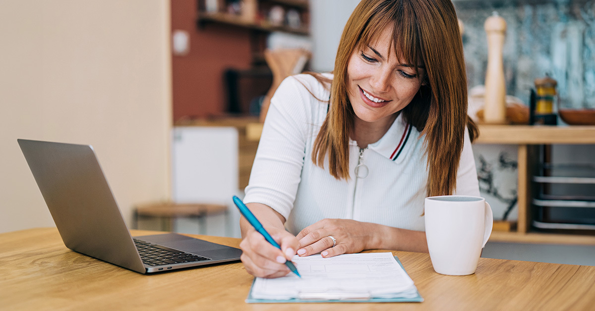 A woman writes on paper at a table with a laptop open in front of her